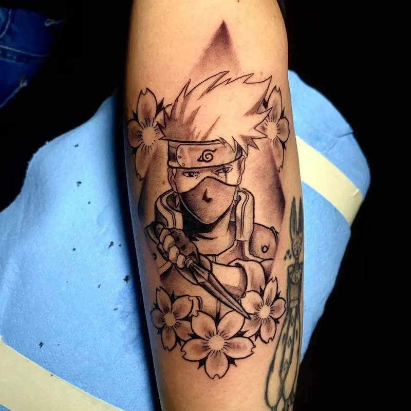 Black and grey anime ninja tattoo on a forearm, showing a masked, headbanded character gripping a kunai knife amid cherry blossoms.