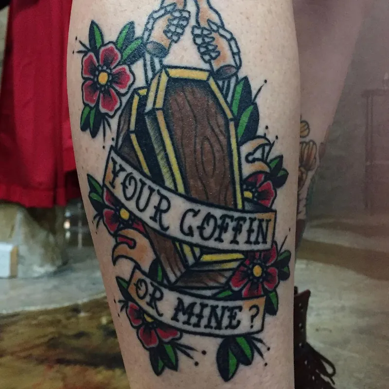Traditional color tattoo on the calf of a coffin held by skeleton hands, framed by red flowers with a banner reading 'Your Coffin or Mine?'