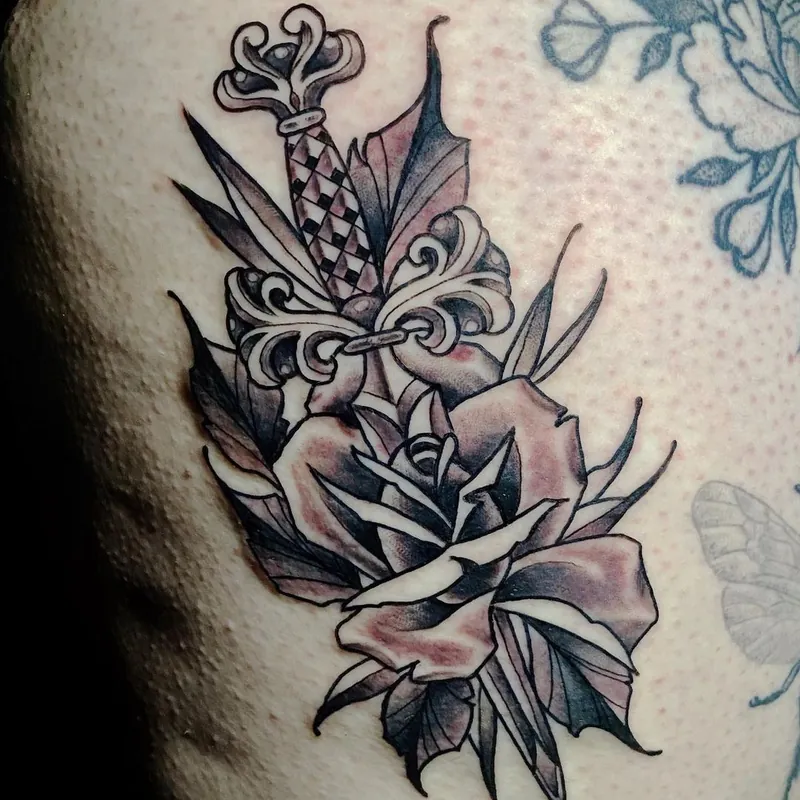 Black and grey neo-traditional tattoo of an ornate dagger with a checkered handle, intertwined with a large rose and leaves.