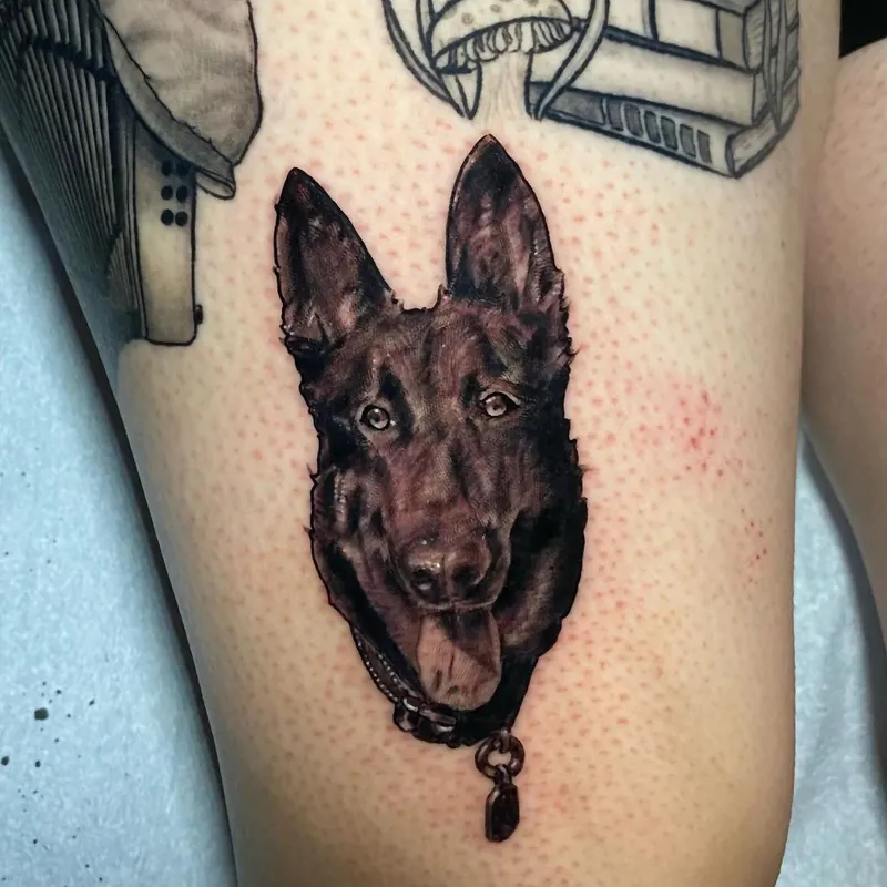 Black and grey realism tattoo on the leg of a dog's face with alert ears, wearing a collar with a hanging charm.