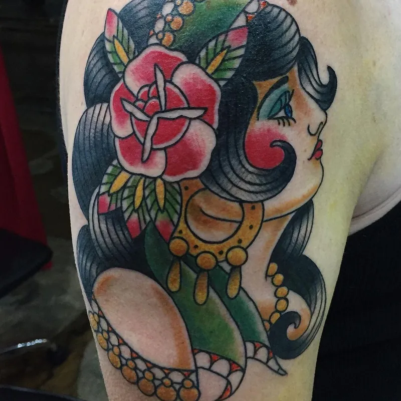 Traditional color tattoo on the shoulder of a woman's portrait in profile, with dark curled hair, a red rose, and gold jewelry.