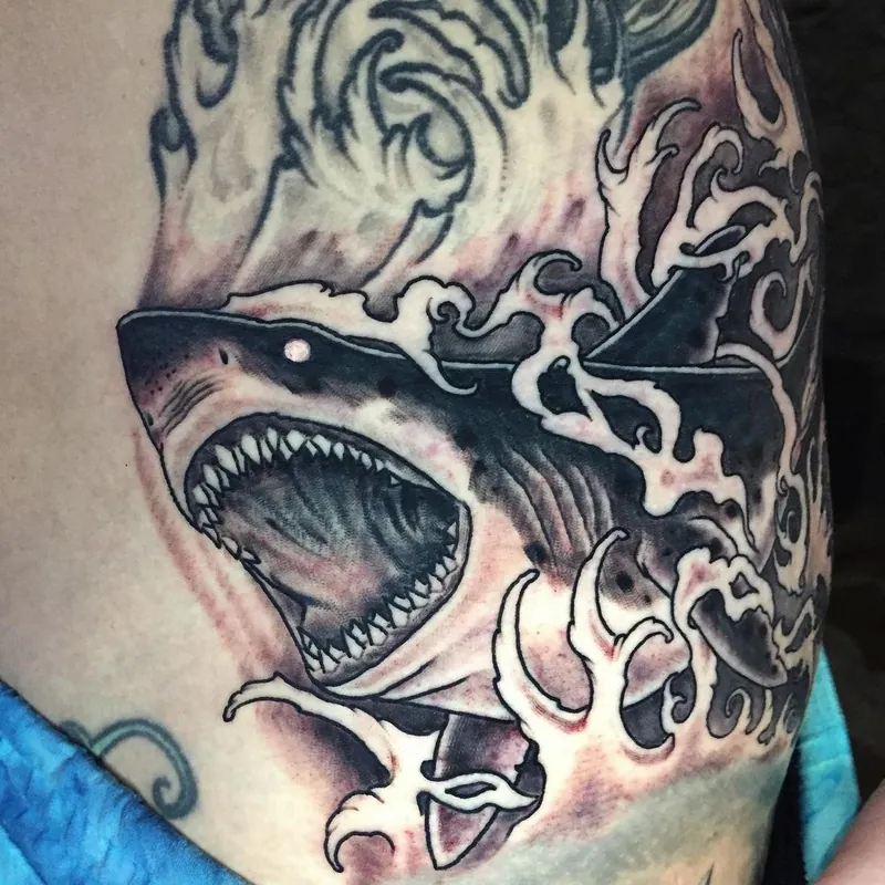 Black and gray tattoo on the side of the torso showing a shark with jaws open baring rows of teeth, swirling Japanese-style waves behind it.