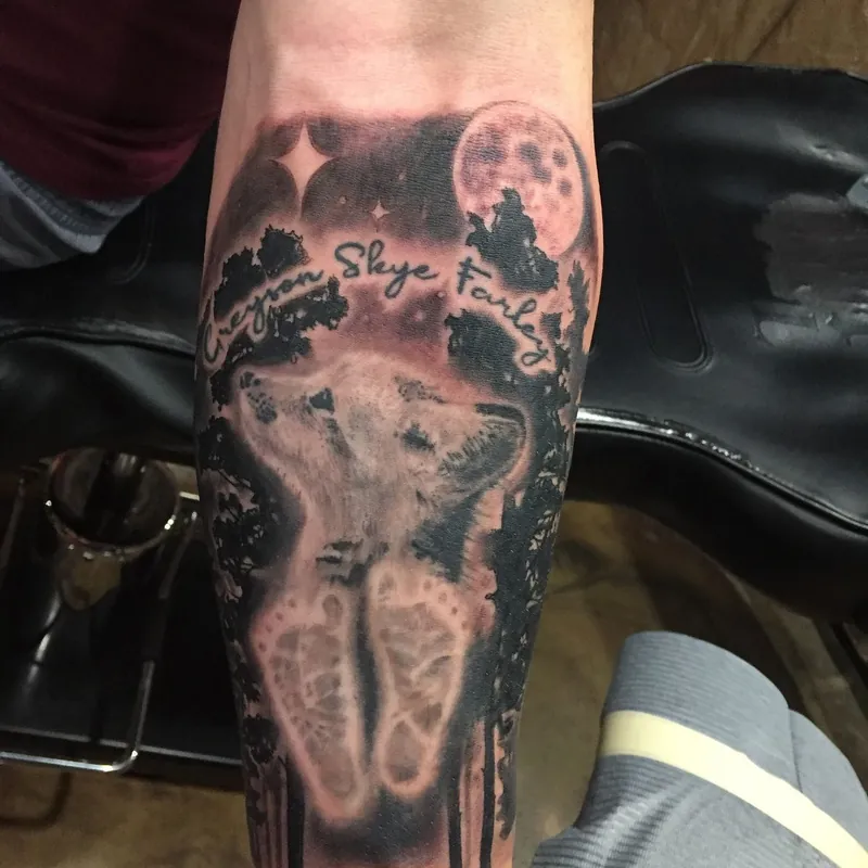 Black and gray memorial tattoo on the forearm featuring a howling wolf beneath a moon, baby footprints, and a name and date in script.