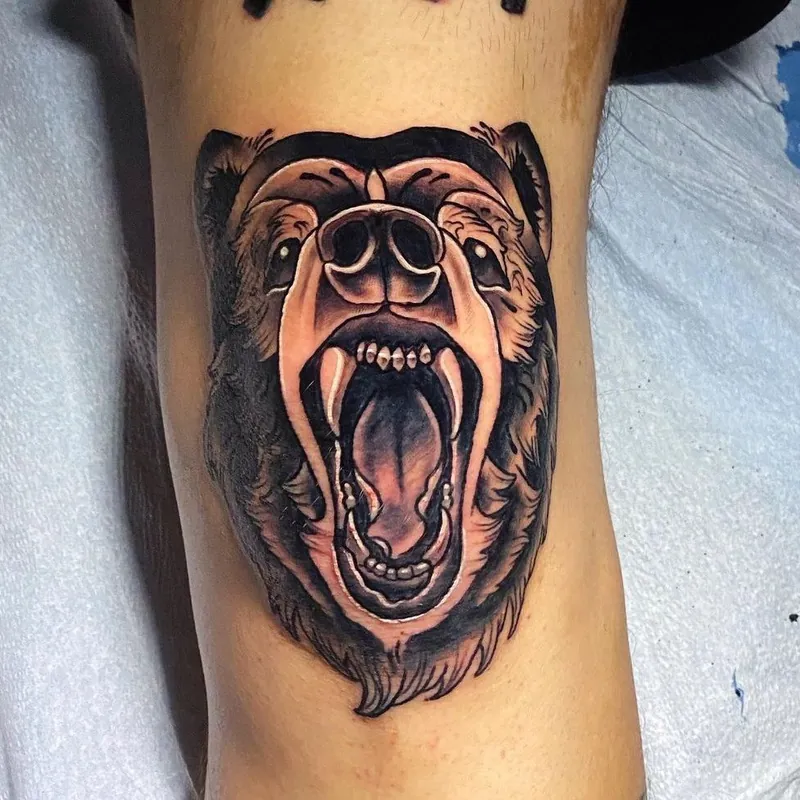 Black and gray tattoo of a roaring bear head with bared teeth and richly textured fur, shaded with warm brown tones.