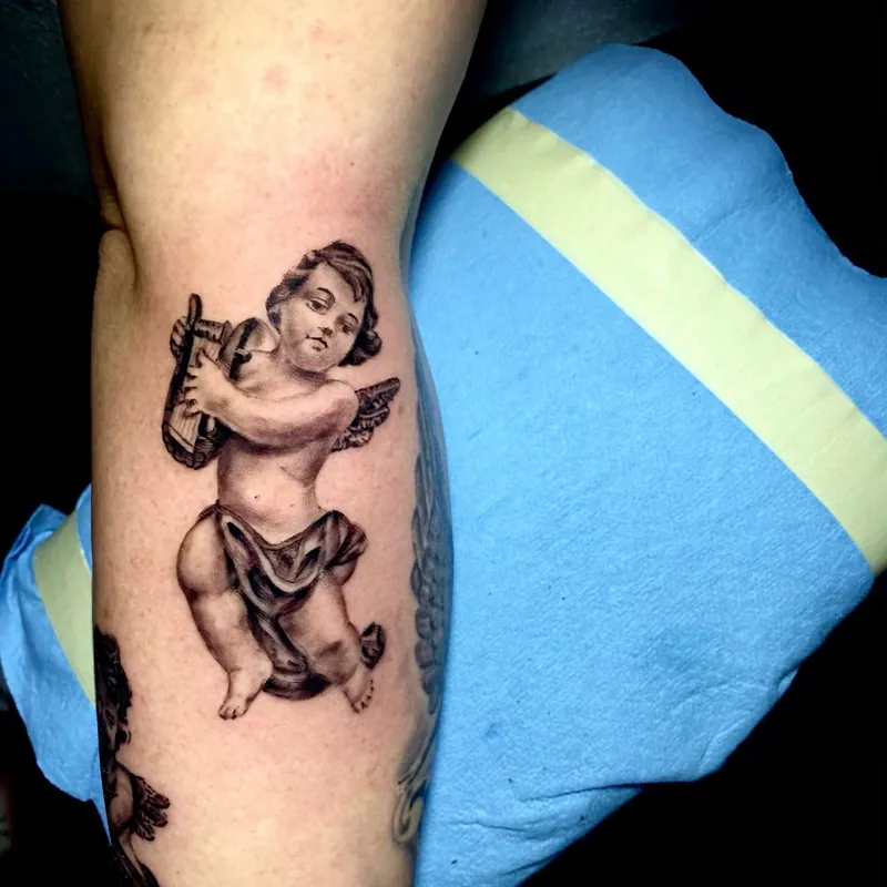 Black and gray tattoo of a cherub playing a small harp, rendered in a classical statue style on the inner arm.