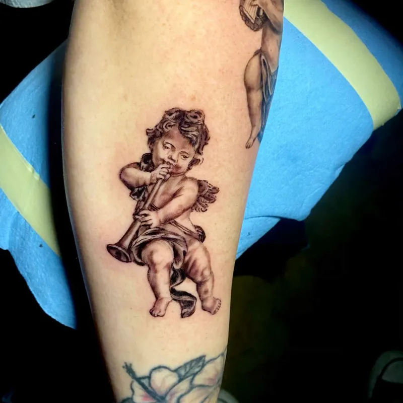 Black and gray tattoo of a cherub playing a wind instrument, rendered in classical statue style on the arm, part of a matching cherub pair.
