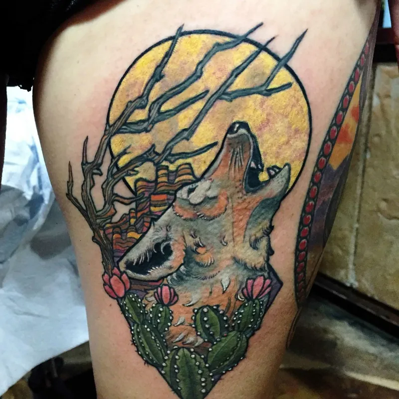 Color tattoo of a howling wolf against a yellow moon with bare branches above and blooming prickly pear cacti below, on the thigh.