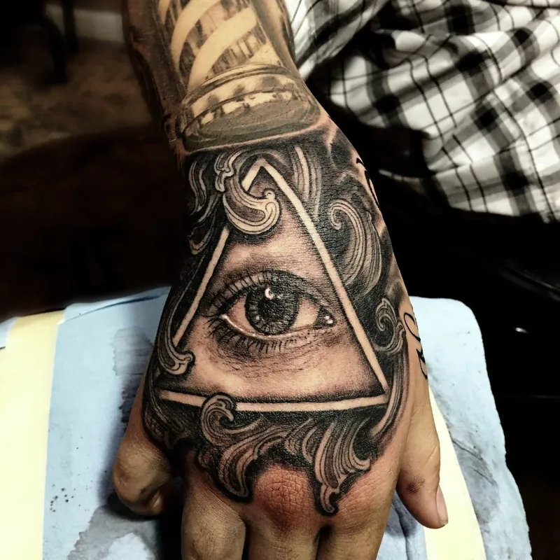 Black and gray hand tattoo featuring the Eye of Providence inside an ornate triangle, with a striped barber-pole design on the thumb.