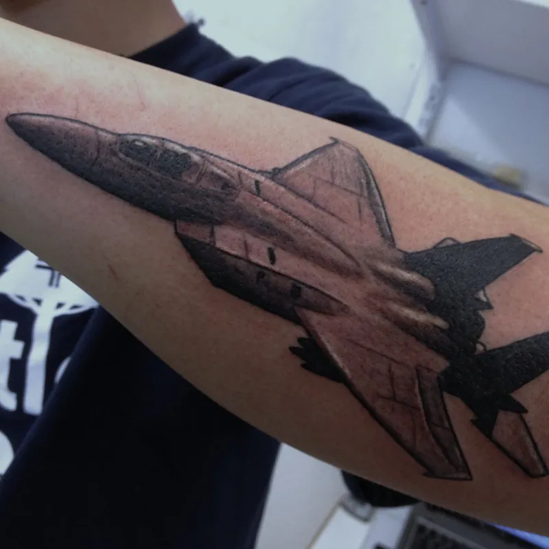 Black and gray tattoo of a fighter jet banking in flight, with swept wings and twin tail fins, on the arm.