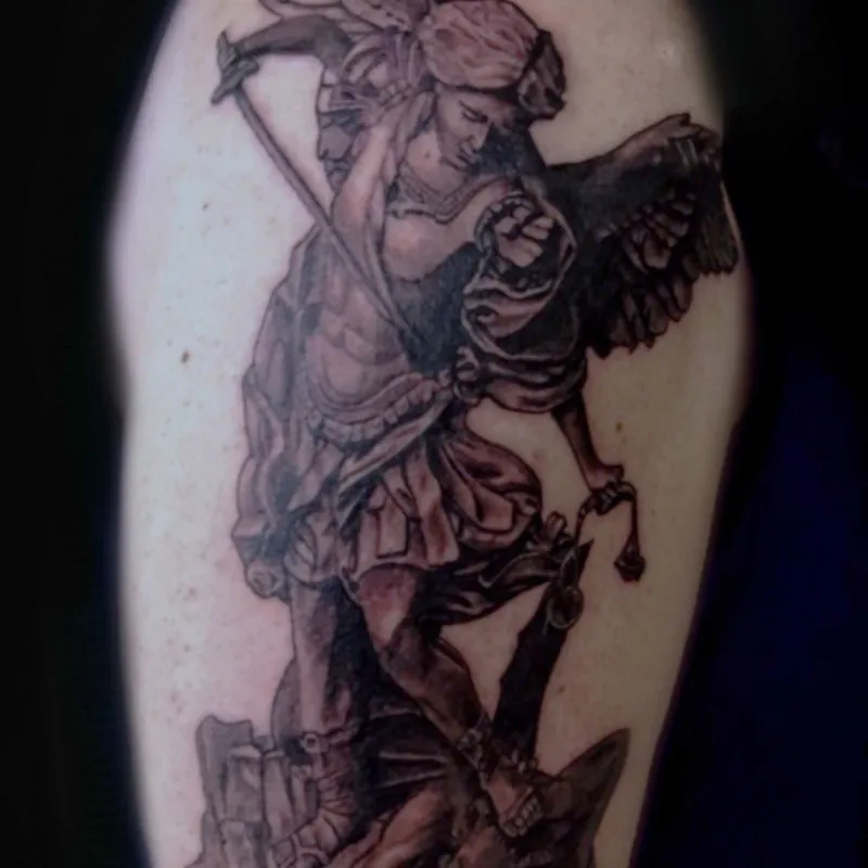 Black and gray tattoo of a winged archangel wielding a sword, standing triumphant over a defeated demon underfoot, in classical statue style on the shoulder.