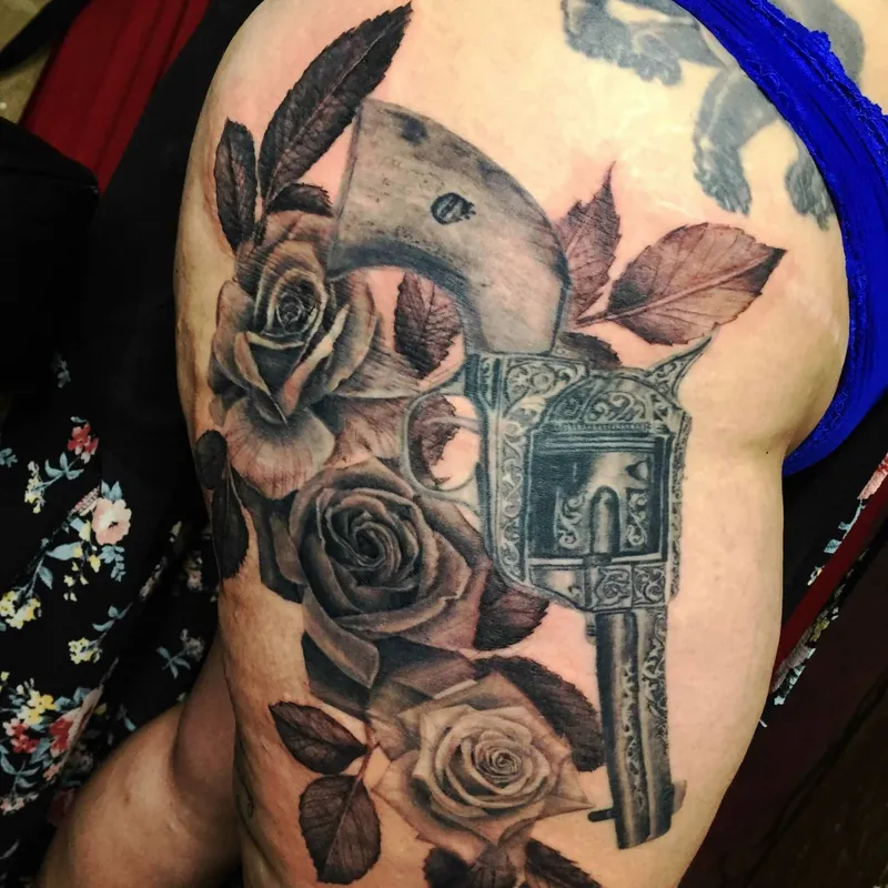 Black and grey realism tattoo of an ornate engraved revolver surrounded by three full-bloom roses, on the shoulder.