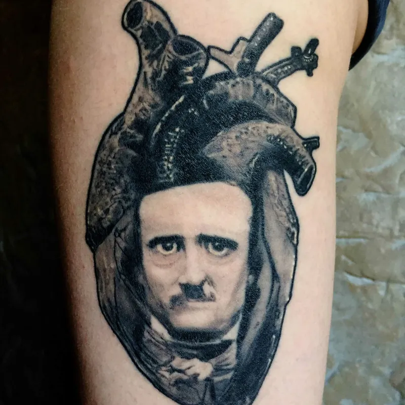 Black and grey tattoo of an anatomical heart with Edgar Allan Poe's portrait forming its lower chamber, on the upper arm.