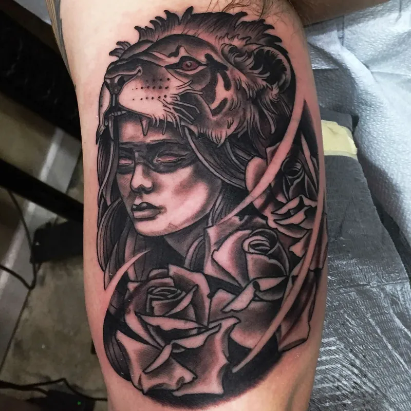 Black and grey neo-traditional tattoo of a woman's face beneath a lion's head and mane, framed by roses on the inner bicep.