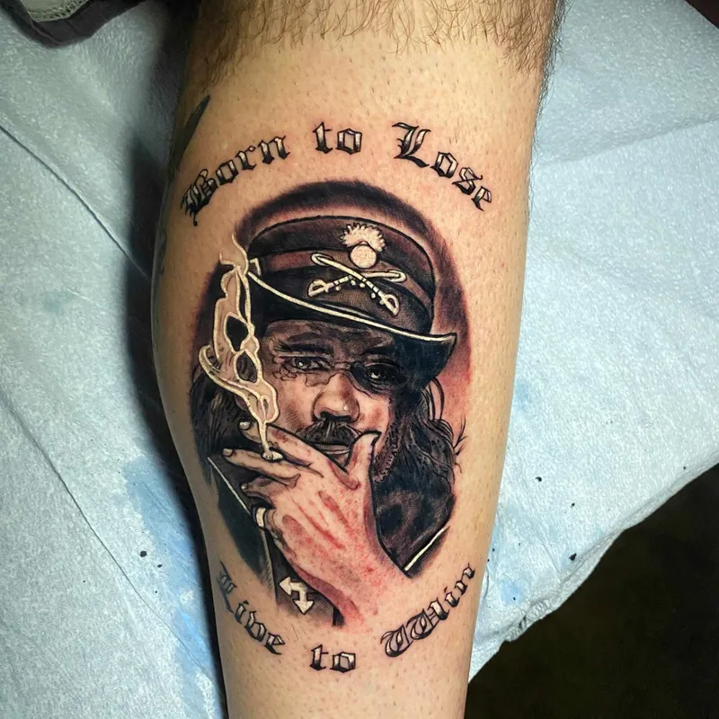 Black and grey tattoo of a man in a peaked cap and eyepatch, framed by 'Born to Lose, Live to Win' script, on the shin.