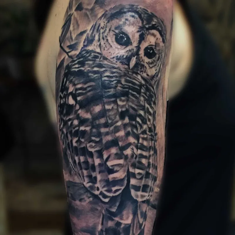 Black and grey realism tattoo of an owl with large dark eyes perched among leaves, on the shoulder.