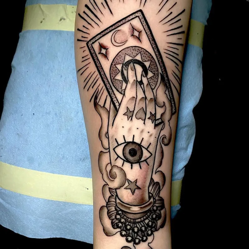 Traditional style tattoo of a mystic's hand holding a moon tarot card, with an eye in the palm and radiating lines, on the forearm.