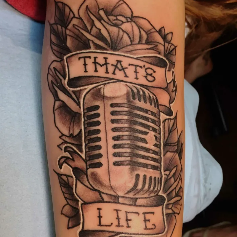 Black and grey traditional tattoo of a vintage microphone surrounded by roses, with banner text reading That's Life, on the forearm.