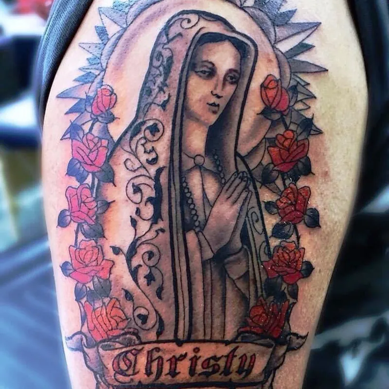 Color tattoo of the Virgin Mary surrounded by red roses, with a banner reading Christy Melany below, on the upper arm.