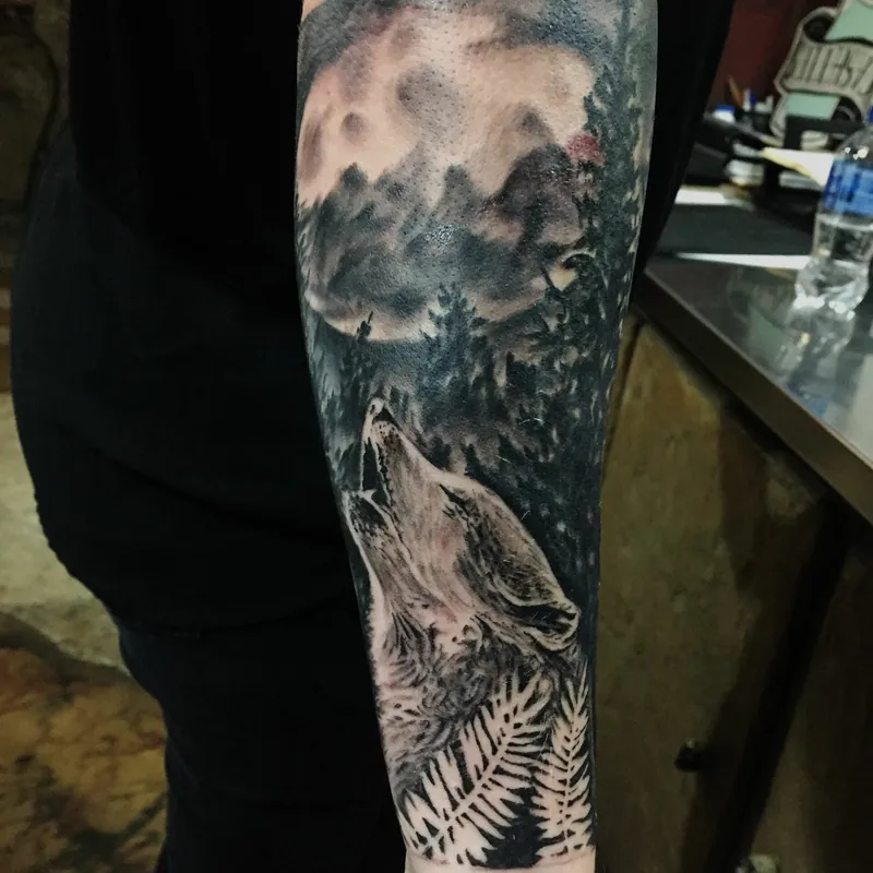 Black and grey realism tattoo of a wolf howling beneath a full moon, with a pine forest silhouette running down the forearm.