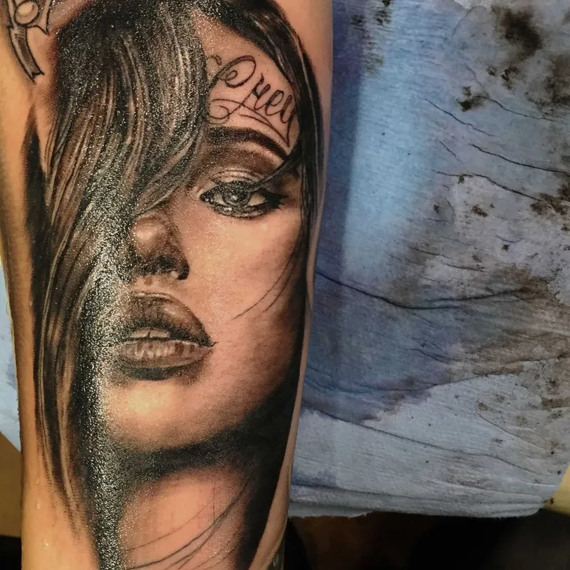 Black and grey realism portrait tattoo of a woman's face with flowing hair falling over one eye and cursive lettering across her forehead, on the arm.
