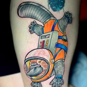 Color tattoo of a platypus character in an astronaut suit, arms and legs spread inside an orange and white spacesuit with blue straps, on the arm.