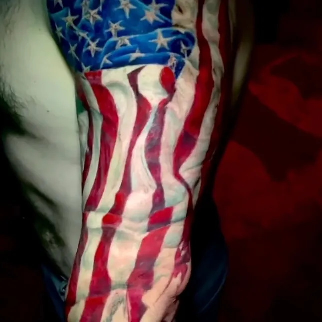 Color tattoo of a tattered, wind-blown American flag running from the shoulder down the arm, with cursive lettering reading the People near the wrist.