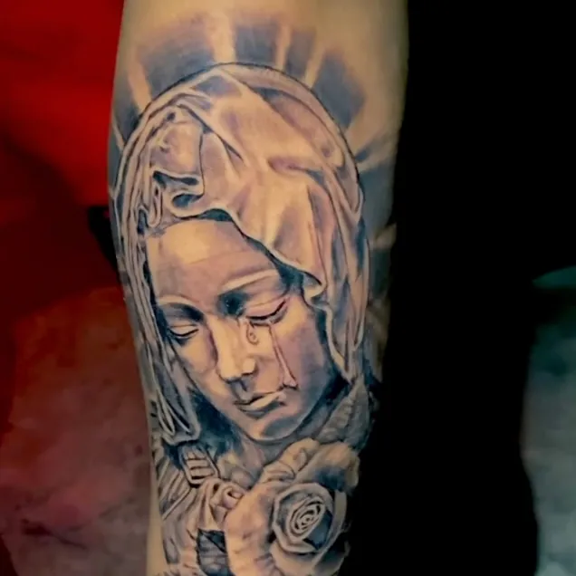 Black and grey tattoo of the Virgin Mary shedding a single tear, haloed by radiating lines, with a rose blooming below on the forearm.