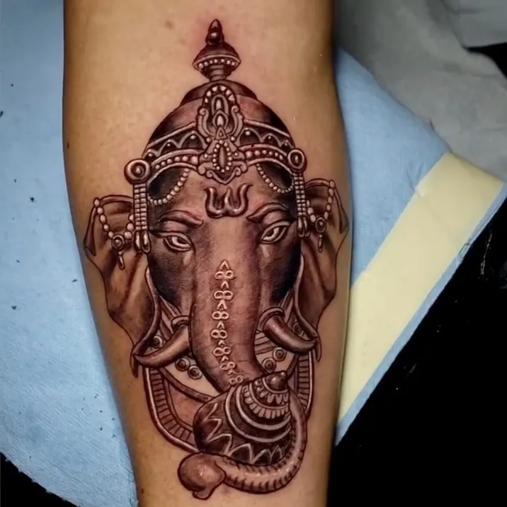 Black and grey realism tattoo of Ganesha's head with an ornate crown, dangling jewelry, and a raised trunk, covering the forearm.