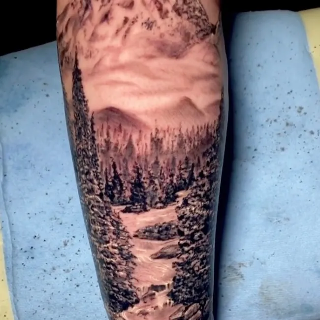 Black and grey realism tattoo of a jagged mountain peak above a pine forest, with a rocky river winding down the forearm.