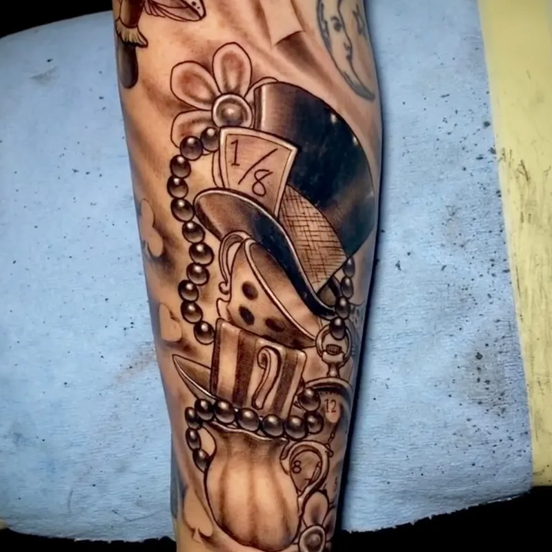 Black and grey neo-traditional tattoo of an Alice in Wonderland scene, with a stacked top hat, teacup, playing cards, and crescent moon on the forearm.