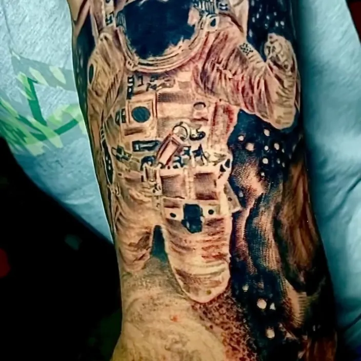 Black and grey realism tattoo of an astronaut in a full spacesuit standing on a lunar surface beneath a starry sky, covering the upper arm.