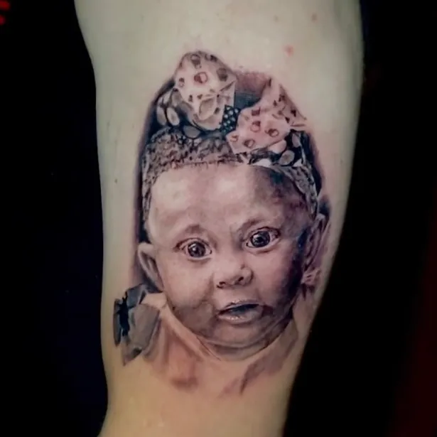 Black and grey realism portrait tattoo of a baby wearing a floral bow headband, finely detailed on the upper arm.