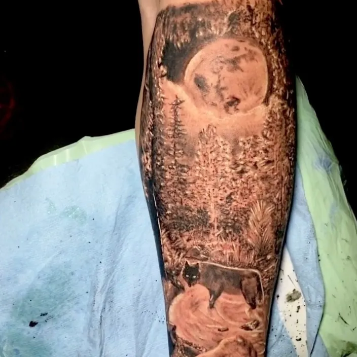 Black and grey realism tattoo of a full moon rising over a pine forest with a wolf below, wrapping down the forearm.