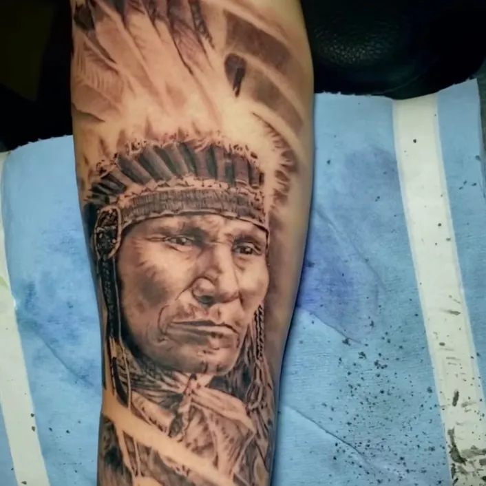 Black and grey realism portrait tattoo of a Native American chief wearing a full feather headdress, on the forearm.