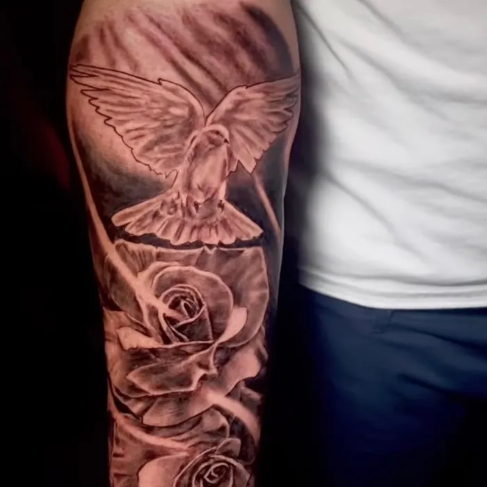 Black and grey realism tattoo of a dove in flight above two roses, with light rays radiating through the design on the forearm.