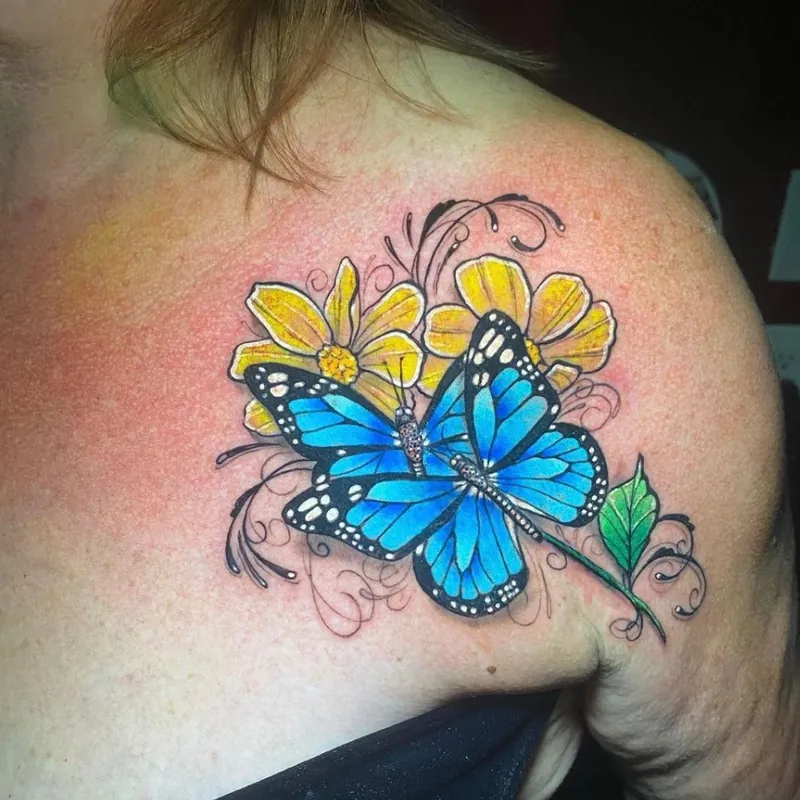 Color tattoo of a blue monarch butterfly resting among yellow daisies with delicate swirl line accents, on the upper chest and shoulder.