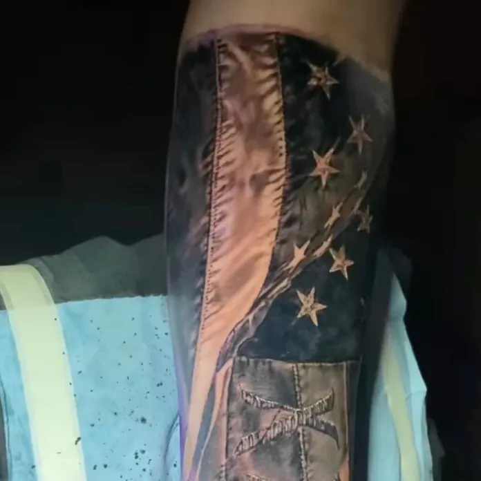 Black and grey tattoo of a draped American flag with detailed fabric stitching and stars, layered above a strapped pouch design on the forearm.