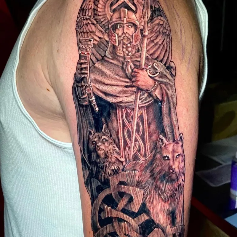 Tattoo of a Norse warrior god in a winged helmet, holding a spear and drinking horn, flanked by two wolves and Celtic knotwork on the upper arm.