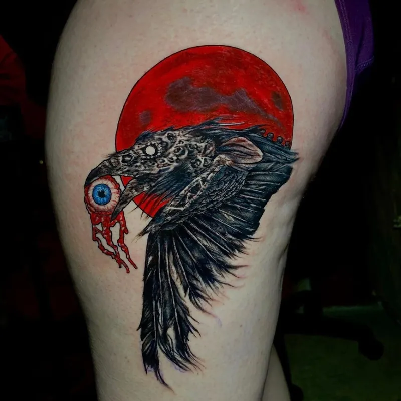 Color realism tattoo of a raven in flight against a red moon, gripping a bloody eyeball in its beak, on the hip.
