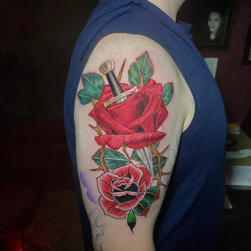 Traditional style tattoo of a red rose pierced by a dagger, paired with a smaller black and red rose below, on the shoulder.