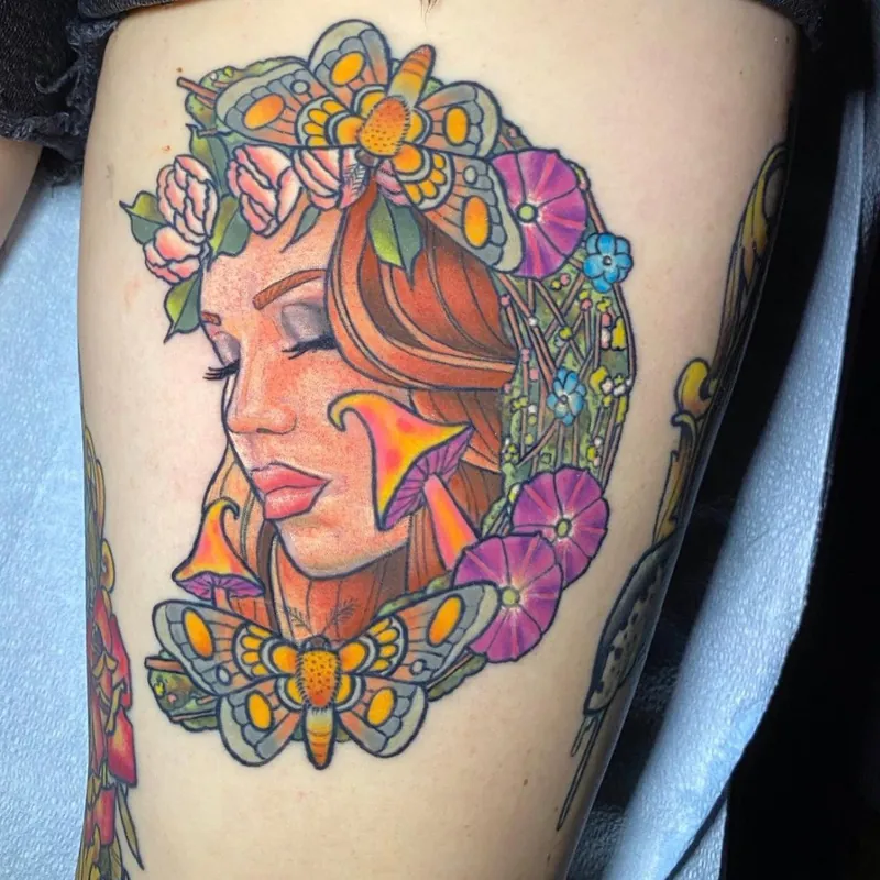 Neo-traditional tattoo of a woman's face with closed eyes surrounded by moths, wildflowers, and mushrooms, on the thigh.