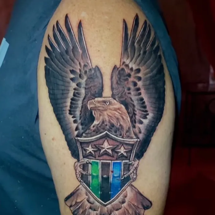 Color tattoo of a bald eagle with wings spread perched above a shield featuring three stars and green and blue stripes, on the shoulder.