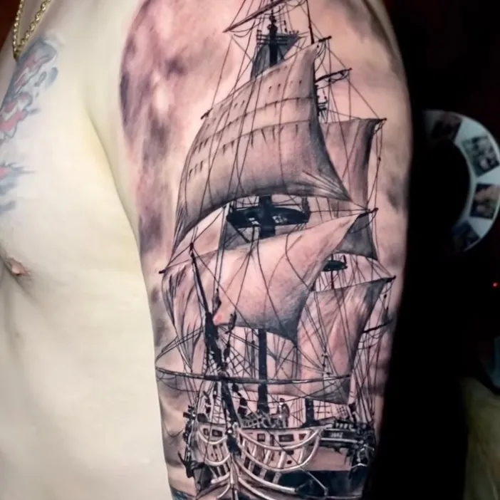 Black and grey realism tattoo of a full-rigged sailing ship with sails unfurled, riding ocean waves, on the upper arm.
