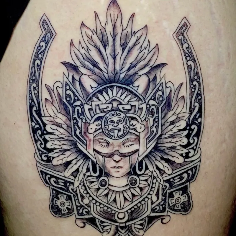 Black and grey fine-line tattoo of an Aztec-style warrior face with closed eyes and an ornate feathered headdress, on the thigh.