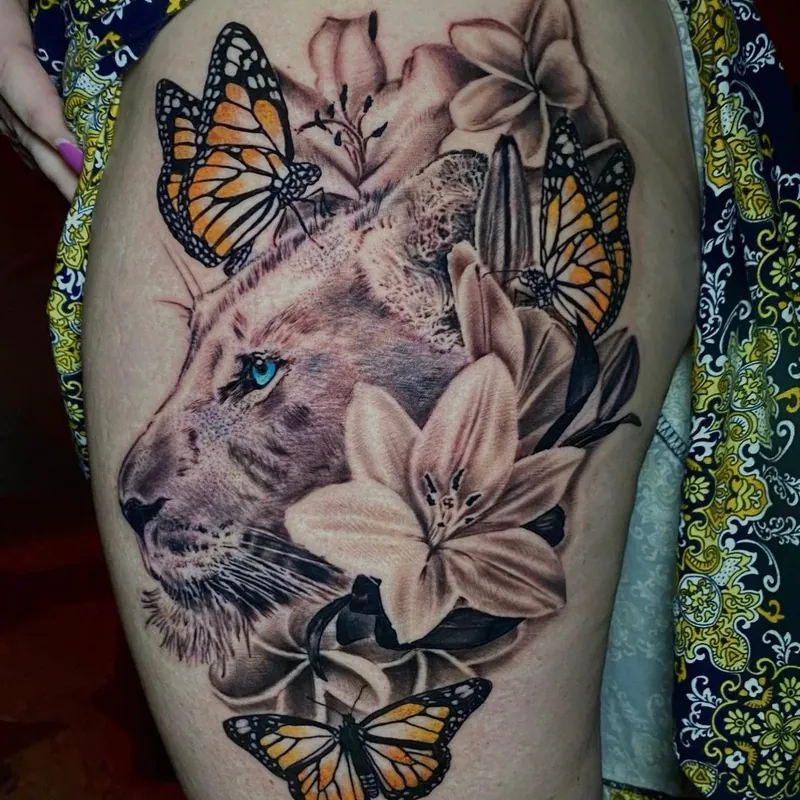Color realism tattoo of a lioness's face with blue eyes surrounded by lilies and monarch butterflies, on the hip.