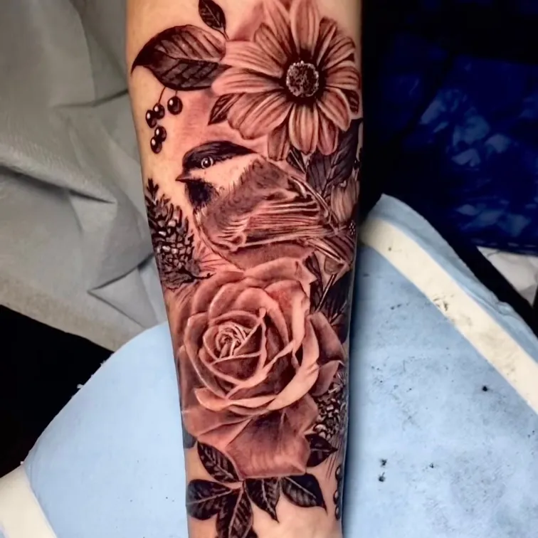 Black and grey realism tattoo of a chickadee perched among a daisy and full rose, on the forearm.
