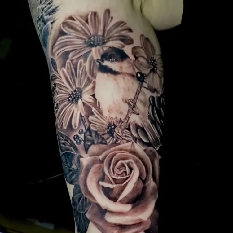 Black and grey realism tattoo of a chickadee among daisies and a large rose, on the upper arm.