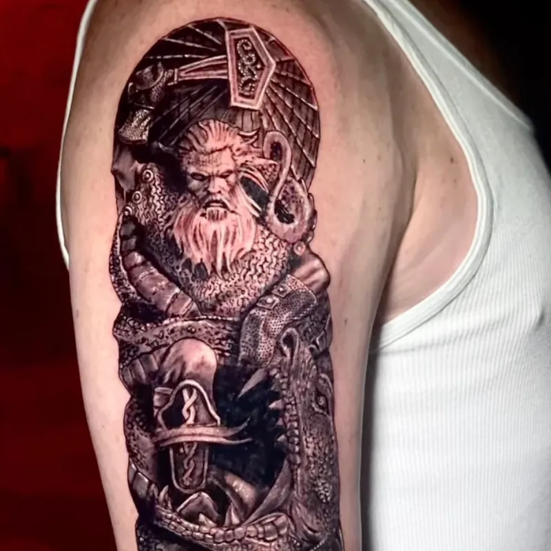 Black and grey tattoo of a horned, bearded warrior entwined in a serpent, seated beneath a rune-carved pendant, above a fanged dragon head, on the upper arm.