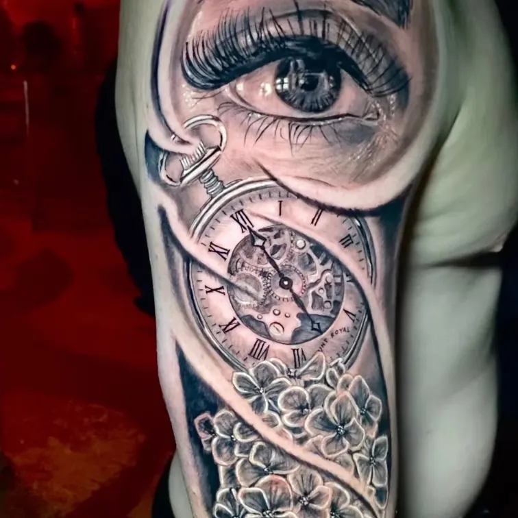 Black and grey realism tattoo of a detailed eye above an open pocket watch with exposed gears, flowing into a cluster of flowers, on the upper arm.