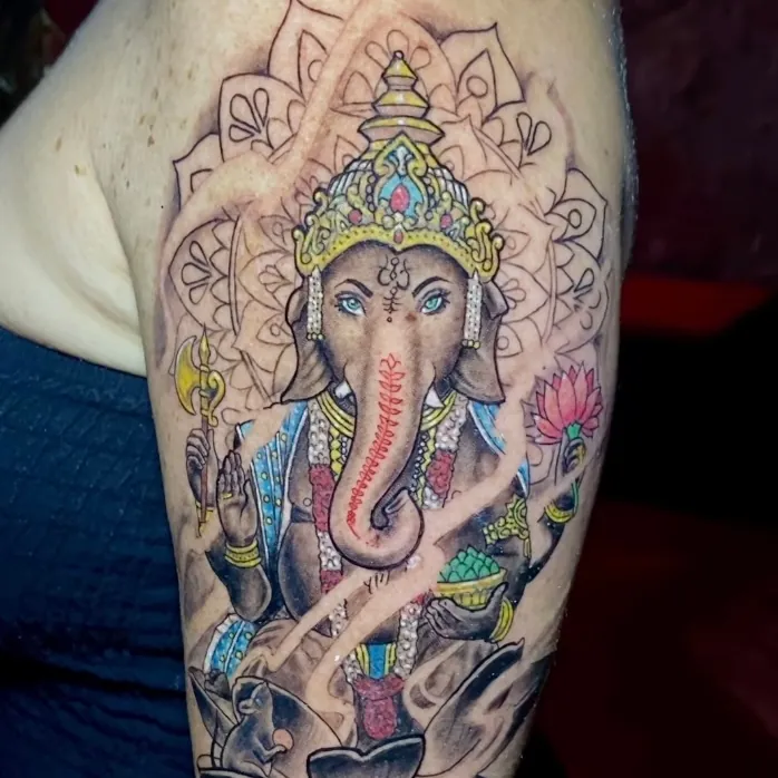 Color tattoo of the Hindu deity Ganesh in an ornate gold headdress, holding a lotus flower and ceremonial items, with his mouse companion below, on the shoulder.